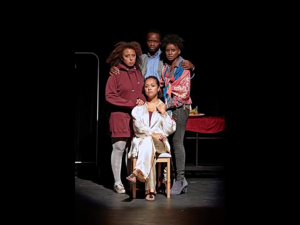 A seated woman surrounded by three figures in An Enemy of the People, staged as a family under civic pressure.