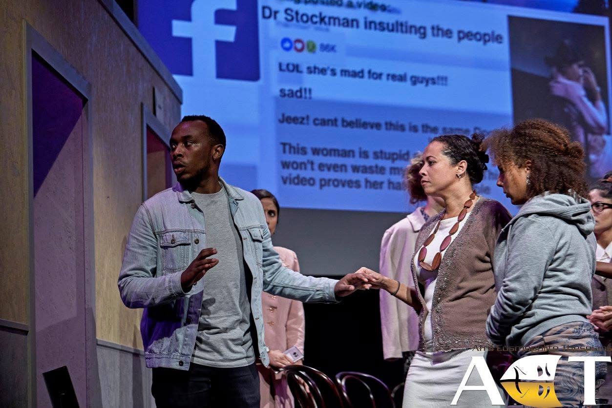 Ensemble scene from An Enemy of the People with social-media projection in the background.